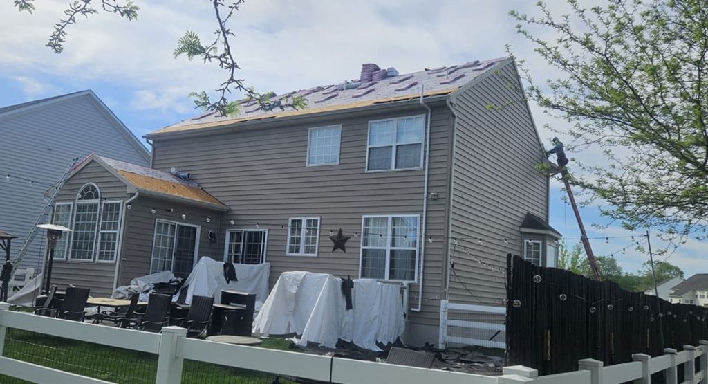 Two-story beige house with roofing work in progress; a worker on a ladder on the right and tarps covering outdoor furniture in the yard.