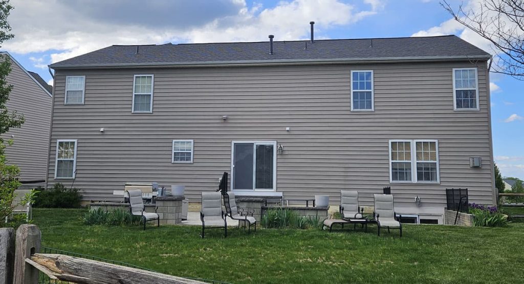Back of a two-story beige house with gray shingles and several windows, a sliding door opens to a yard with outdoor chairs and a stone patio area under a partly cloudy sky.