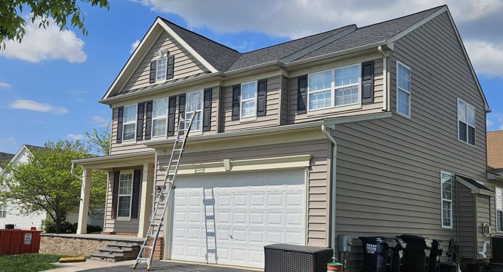 Two-story beige suburban home with a ladder resting against the garage door and several black trash bins nearby under a blue sky.