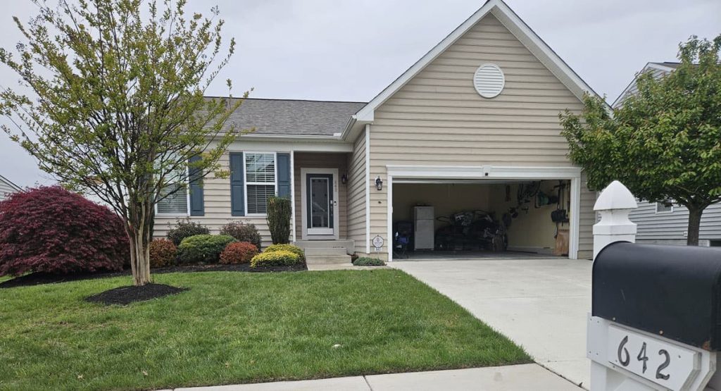 Front view of a beige single-story house with an open two-car garage cluttered with belongings, a manicured lawn, and trimmed shrubs.