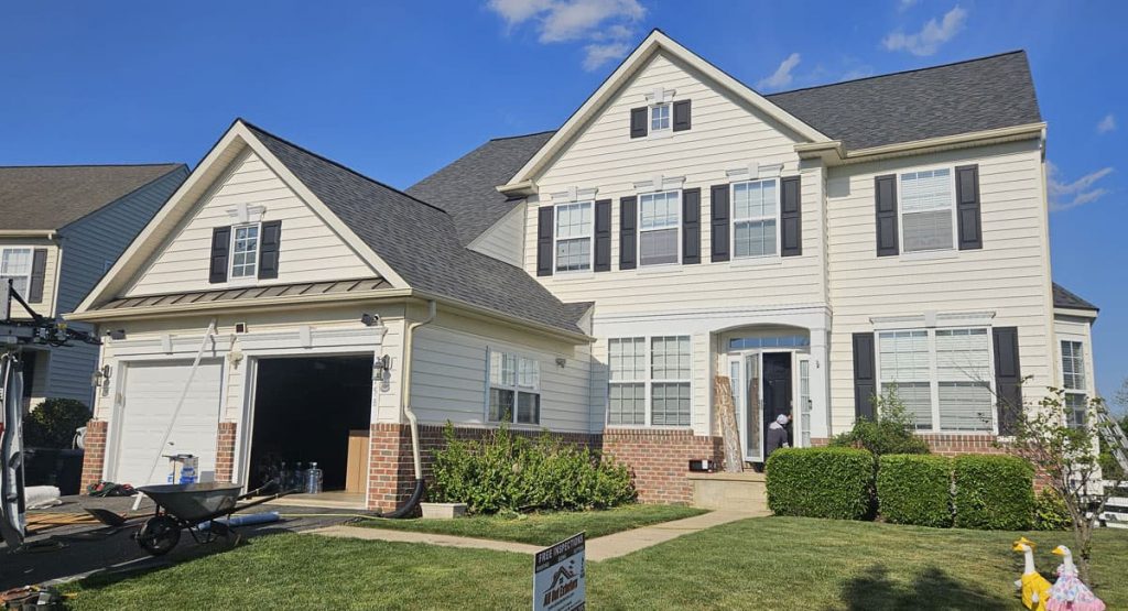 Two-story suburban house under renovation with an open garage, construction materials, and a worker at the front door on a sunny day, with a wheelbarrow nearby.