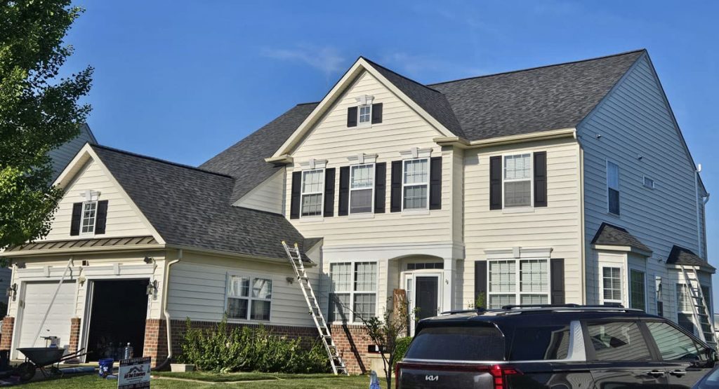 Two-story beige house with black shutters undergoing exterior renovation; ladders lean against the front, with tools and a sign in the yard.