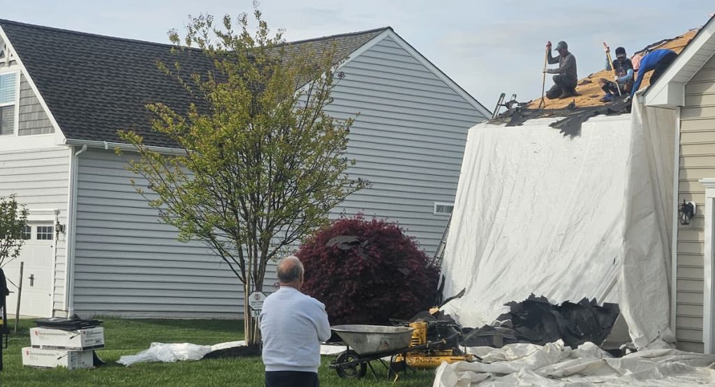 Roof workers secure a large white tarp over a house roof while a man watches from the yard beside a wheelbarrow and construction supplies.
