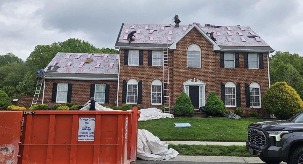 Roofing crew on the two-story brick house installing pink underlayment, with an orange dumpster and a truck in the driveway.