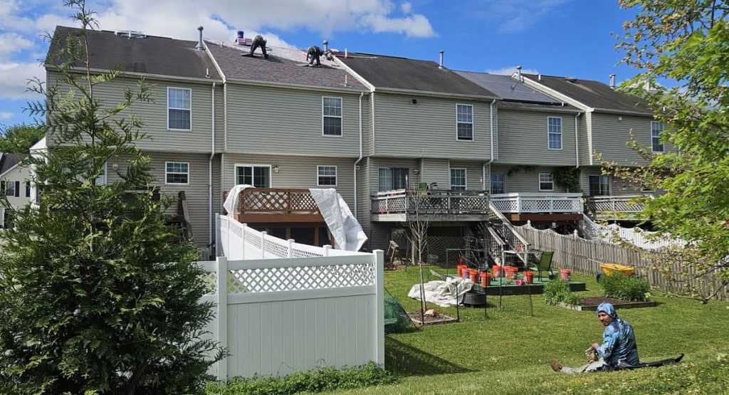 Row of townhouses with workers on the roofs, a white fenced yard in the foreground, and a person in a blue headscarf sitting on the lawn.