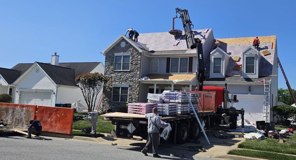 Roofing crew on a two-story suburban house, crane lifting materials onto the roof with pallets stacked on a flatbed truck nearby.