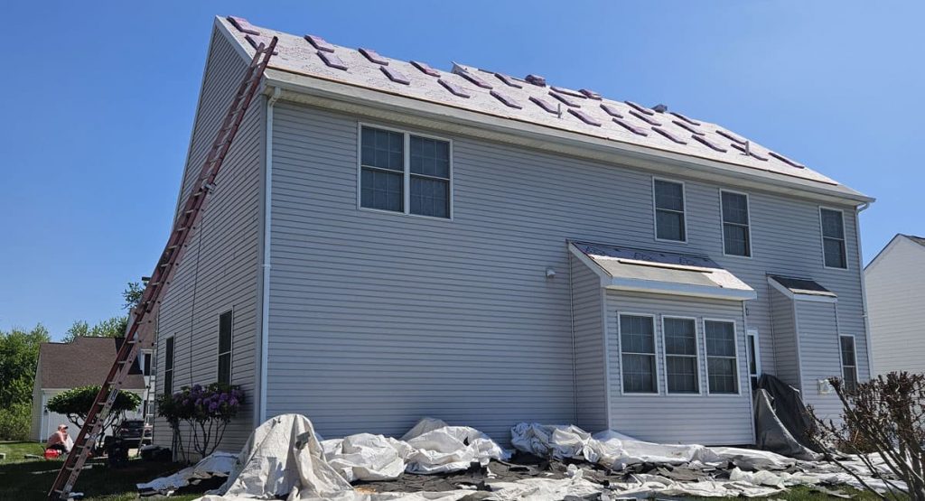 Two-story suburban house with roof under construction, a ladder leaning against the left side and tarps noisily spread on the ground.