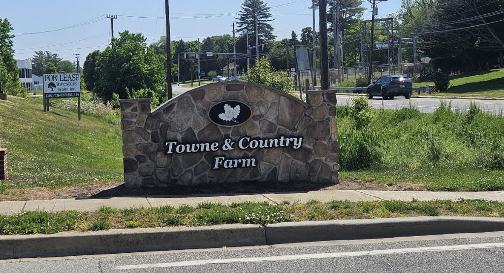 Stone sign reading 'Towne & Country Farm' with a leaf emblem, set along a road with greenery and a 'For Lease' sign nearby.