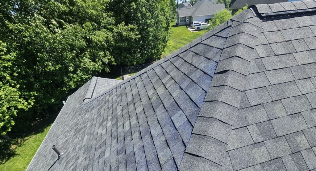 Aerial view of a dark gray shingled roof with neighboring houses and green trees in the background.