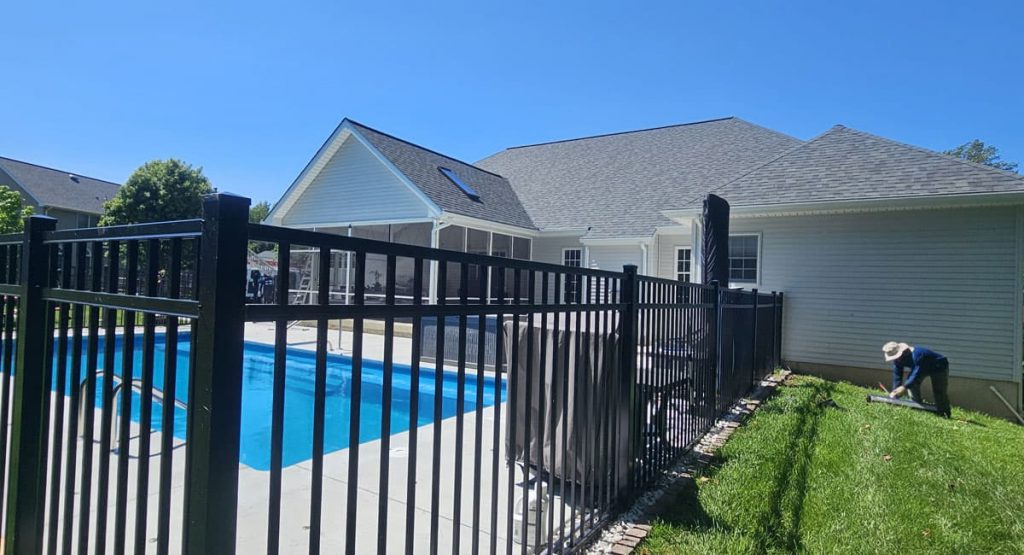 Backyard with a blue in-ground pool behind a black metal fence and a worker trimming grass near the house on a sunny day.