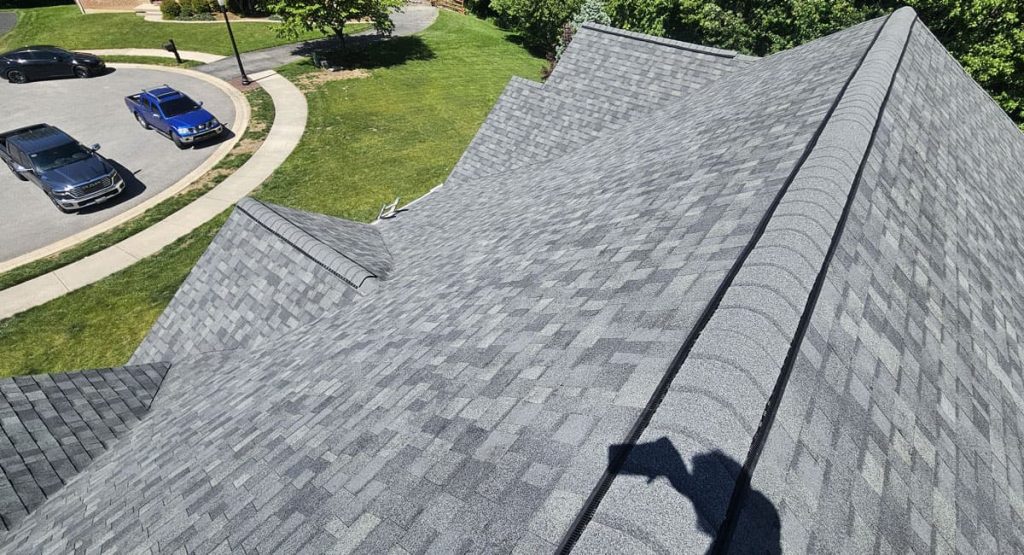 Aerial view of a grey shingled roof with multiple peaks, adjacent curved driveway, and parked cars on a grassy cul-de-sac.