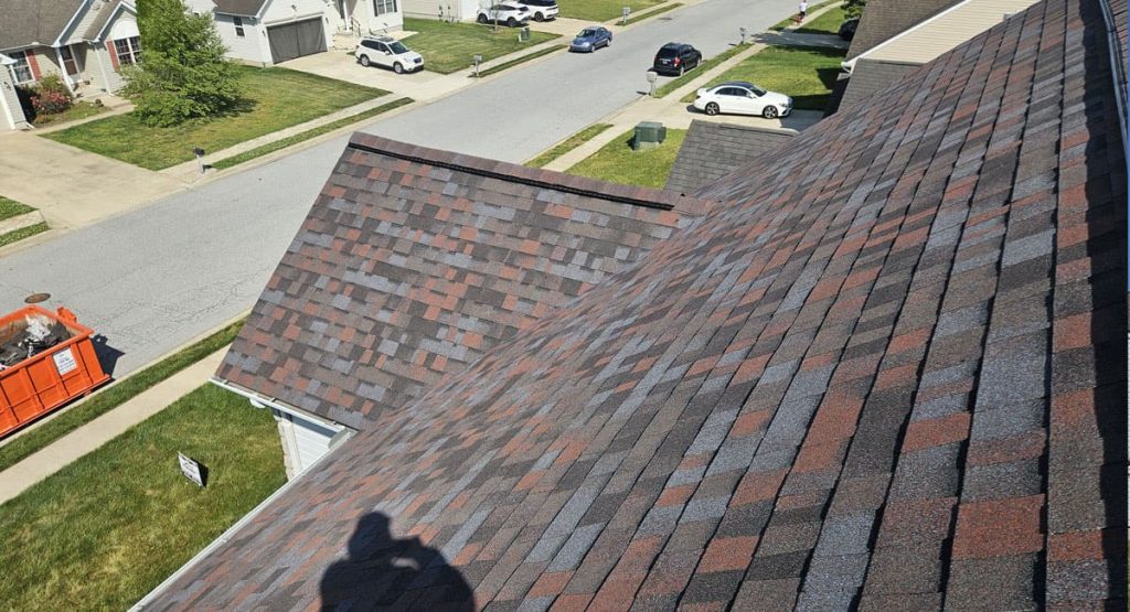 Aerial view of a suburban neighborhood showing a house roof with dark brown and gray shingles, plus a red dumpster on the left lawn.
