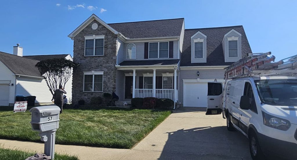 Two-story suburban house with a stone facade on the left and a white porch, with a ladder-equipped van parked in the driveway and workers in the yard.