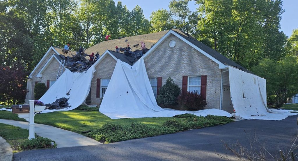 House with extensive roof tarps covering damaged shingles after a storm, workers on the roof aiding repairs in a suburban yard.