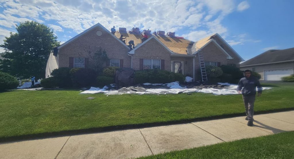 House with workers on the roof during roof repair; tarps and debris spread across the lawn, ladder propped at the front door.
