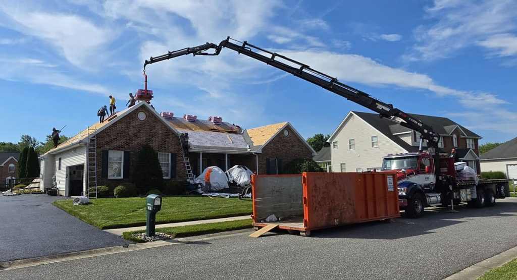Crew on a brick house roof install new shingles while a crane lifts materials; an orange dumpster sits in the street nearby.