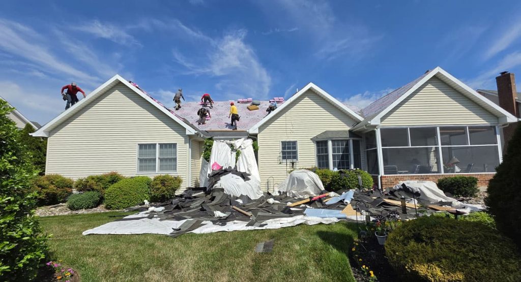 Suburban house undergoing roof replacement with workers on the pitched roof and torn shingles scattered across the front lawn.