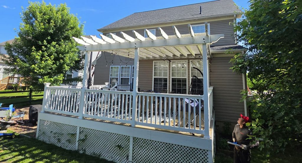 White wooden deck with a pergola attached to a beige house, with a person in a red headband standing nearby in the yard.