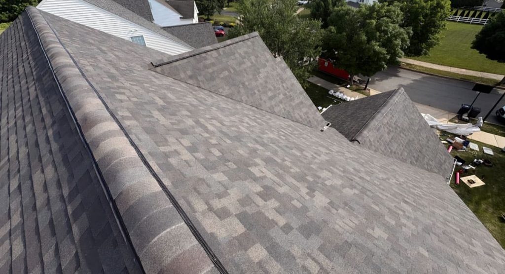 Aerial view of gray shingle roof with multiple steep gables; construction materials visible in a yard nearby.