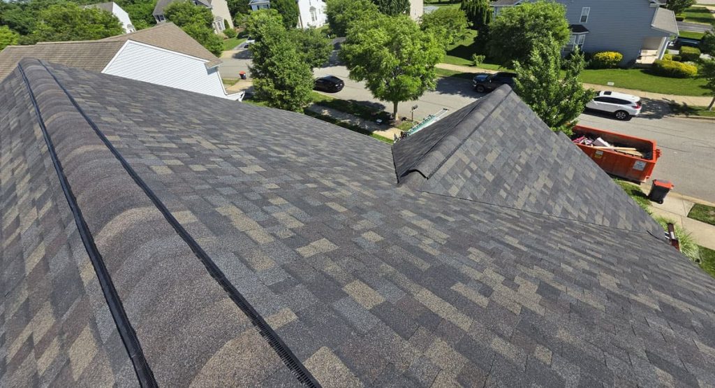 Aerial view of a large gray shingled roof in a suburban neighborhood, with trees, parked cars, and a dumpster on the street nearby chevroned by roofs.
