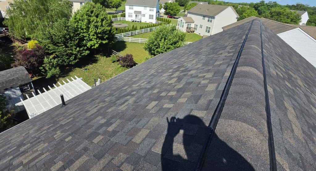 Aerial view of a sloped, dark gray shingled roof with a long ridge vent, shadow of the photographer, and green trees and houses in a suburban yard below.