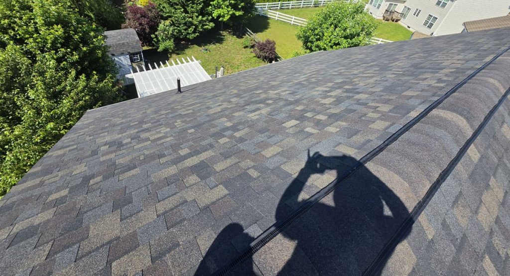 Aerial view from a pitched roof showing gray asphalt shingles and surrounding yard with trees and a white fence; photographer's shadow on the roof.