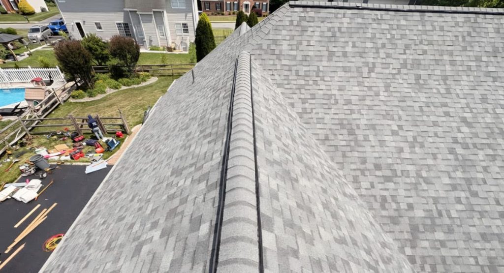 Aerial view of a light-gray shingle roof with a central ridge, neighboring yard with construction items and tools nearby