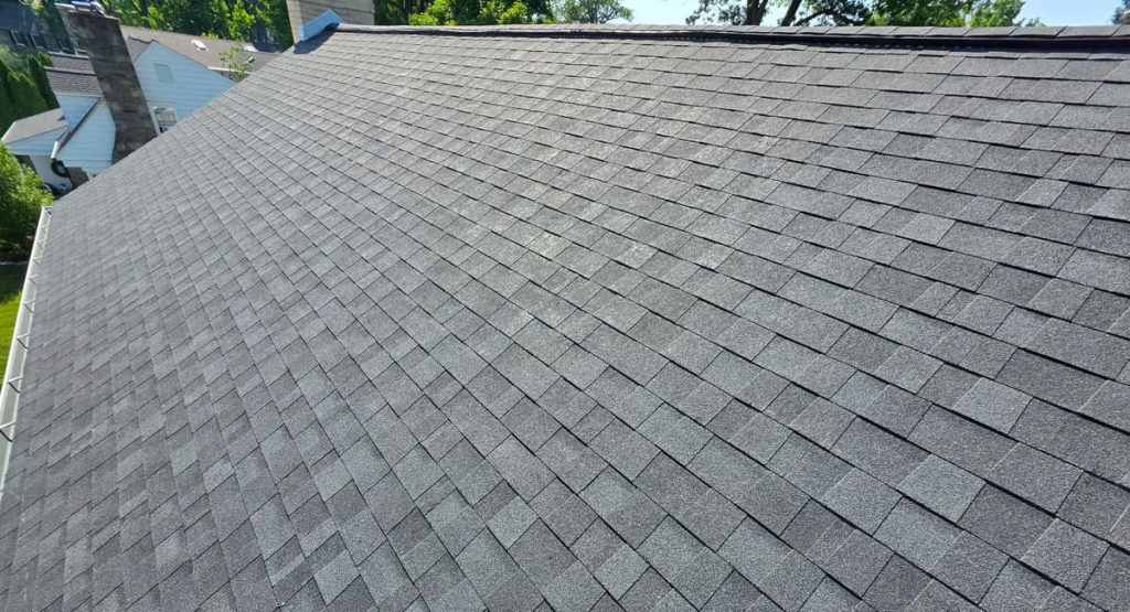 Aerial view of a gray asphalt shingle roof sloping downward with trees and neighboring houses in the background.