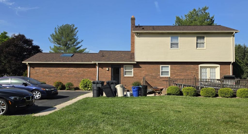Front view of a two-story brick-and-beige suburban house with a well-kept lawn and a row of trash and recycling bins in the driveway.