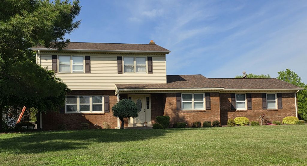 Two-story suburban house with a brick lower level, beige siding on the upper level, and brown shutters, front lawn and shrubs nearby.