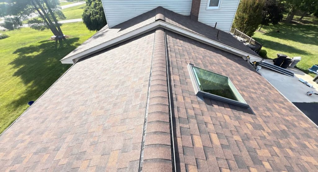 Aerial view of a brown shingle roof with a skylight, chimney, and surrounding green yard.