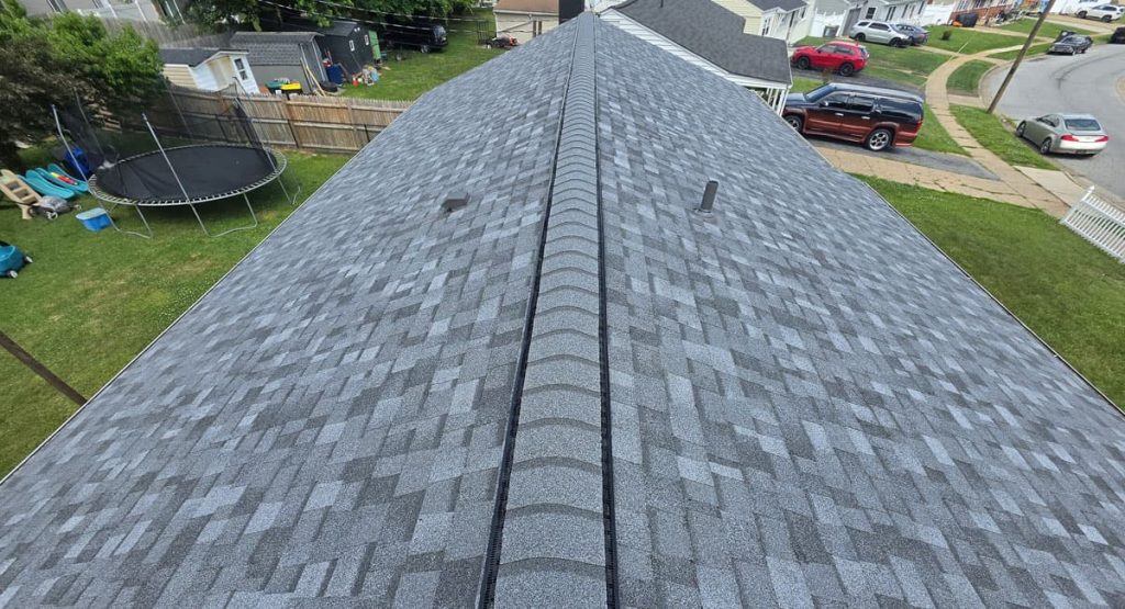 Aerial view of a grey shingled roof with a central ridge vent, neighborhood houses and a trampoline visible in the yard nearby.