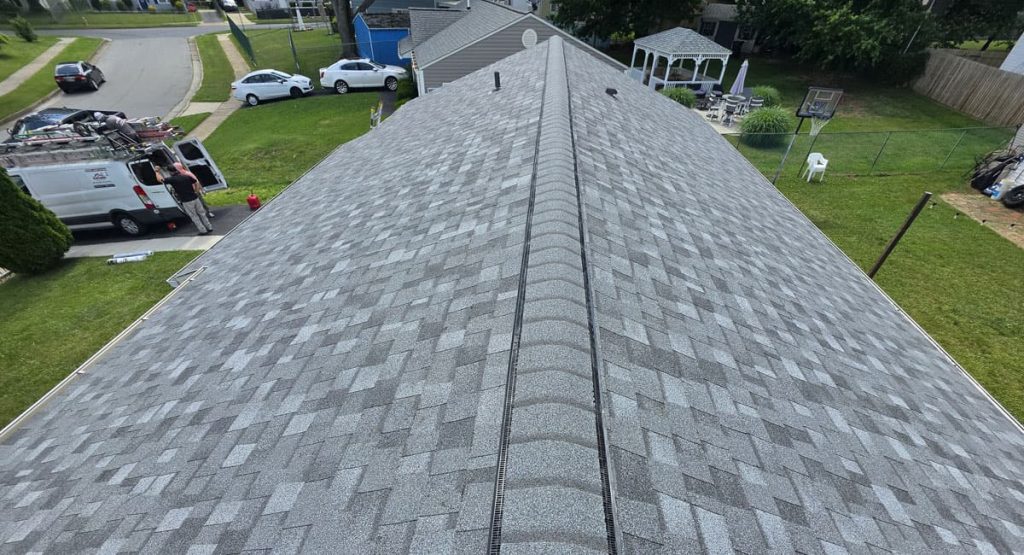 Aerial view of a gray shingle roof with a central vent seam; a white van with workers and equipment on the left, in a suburban neighborhood.