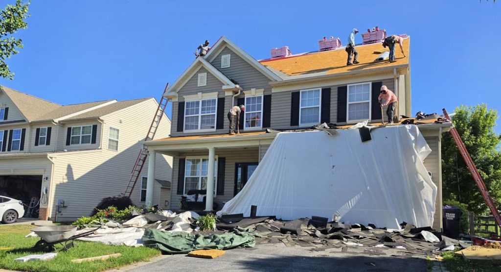 Two-story suburban home with the roof being replaced; debris and tarps cover the driveway in front of the house.