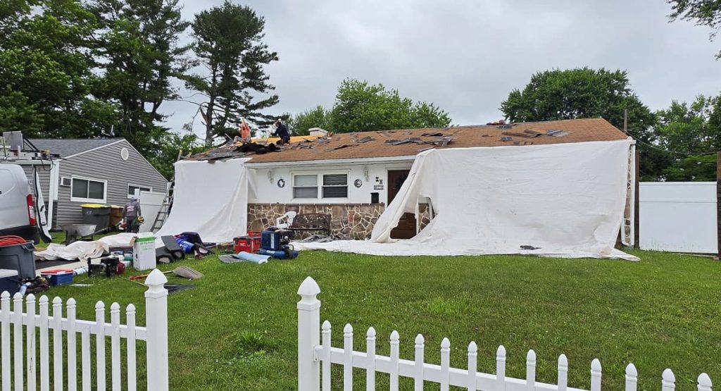 Front view of a single-story home being reroofed after a storm; two workers on the roof, missing shingles, and tarps covering side areas with tools and debris in the yard, white picket fence in the foreground.