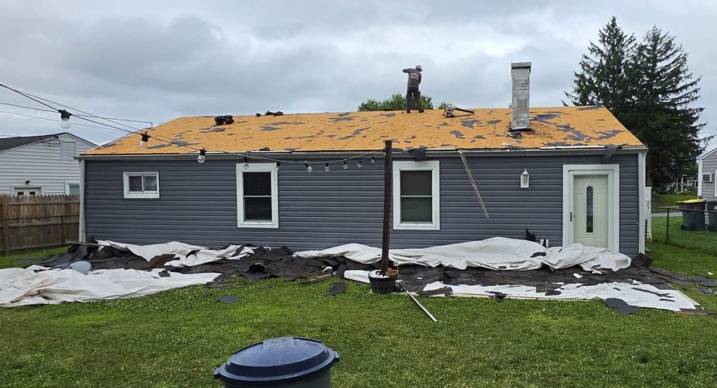 Worker on a blue, single-story house roof under renovation with torn orange underlayment; large white/black tarps and debris scattered in the yard behind the house.