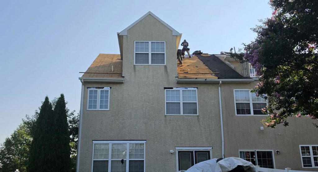 Two workers on a beige multi-story house roof under renovation, missing shingles and plywood exposed against a clear blue sky.
