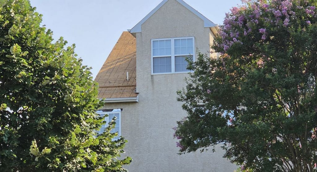 Two-story beige house with a peaked roof, visible white-framed window, framed by green trees on both sides.
