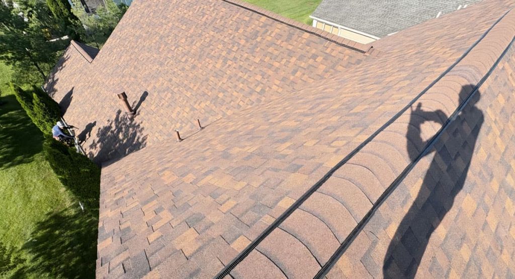Aerial view of a brown shingle roof with a worker on a ladder trimming a hedge near vents; photographer's shadow visible on the roof.