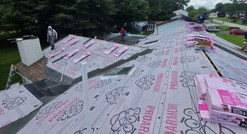 Workers installing roof underlayment on a sloped house with pink insulation bundles nearby and a ladder visible on the left.