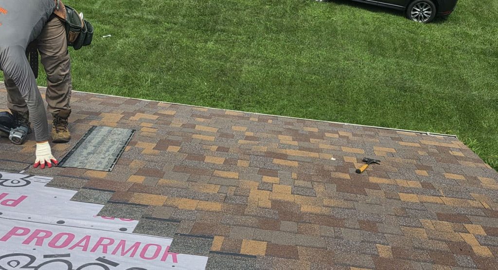 Person installing roof shingles on a sloped roof; one hand rests on a metal starter strip while tools lie nearby and a roll of underlayment shows pink text at the edge of the roof surface.