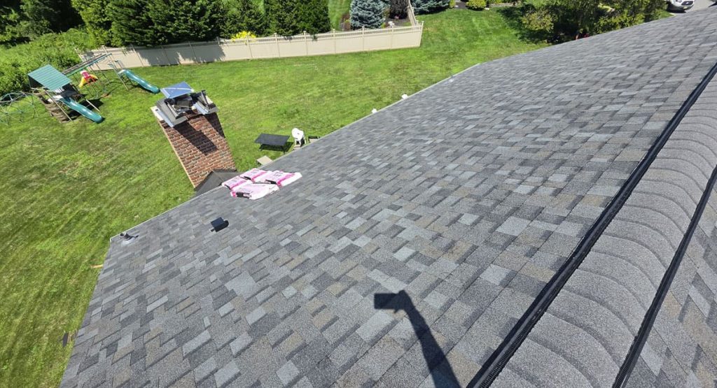 Aerial view of a gray shingled roof with a backyard and playground beyond, bags and tools on the roof, and the photographer's shadow visible on the shingles.