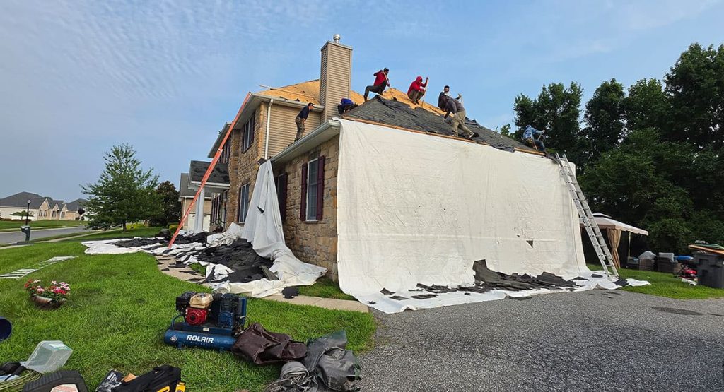 House undergoing roof replacement; workers on the roof, white tarps covering the wall, and tools/ladder in the yard ahead of the home renovation.