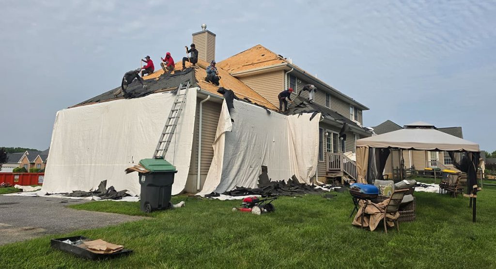 Roofing crew works on the roof of a damaged two-story house while tarps cover the walls and debris lies in the yard a ladder leans against the roof.