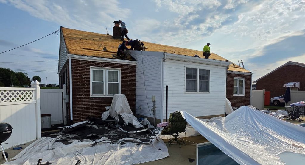 Workers on the roof of a small brick-and-siding house repairing shingles, with roofing debris and white tarps scattered in the yard under a sunny sky