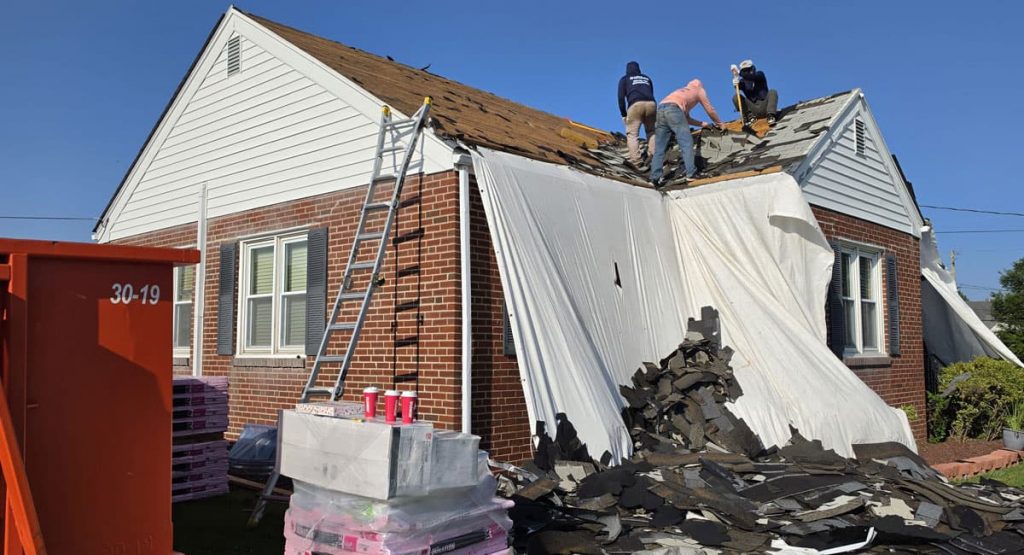Roofers remove shingles from a pitched roof with a ladder against a brick house and white tarp covering part of the wall; blue sky above.