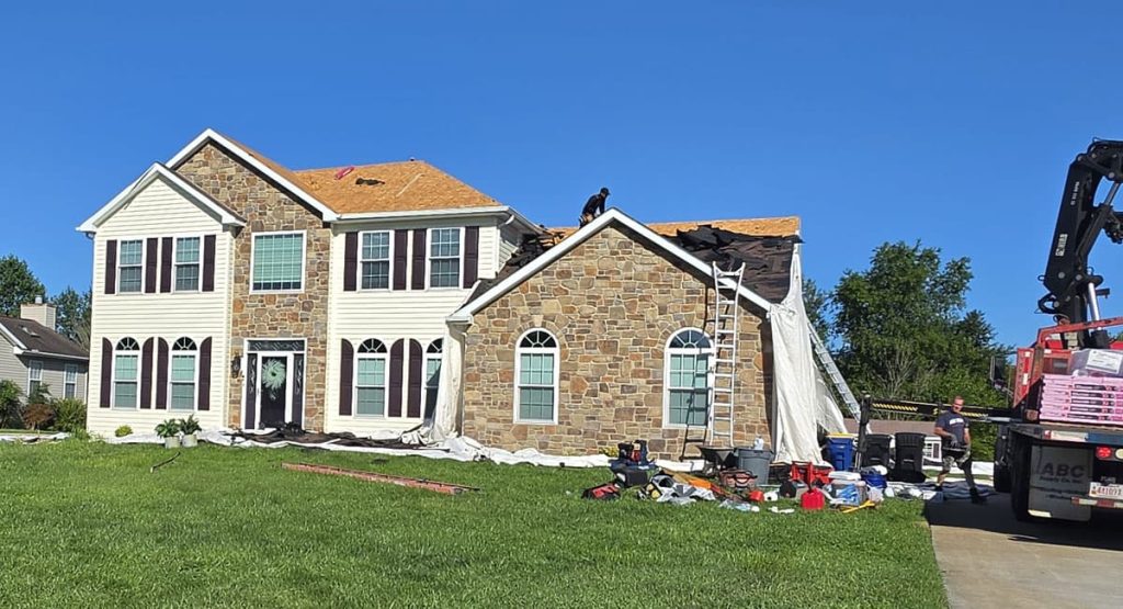 Front view of a suburban house under roof repair, with a ladder on the stone facade and workers on the roof amid tarps and tools on the lawn.