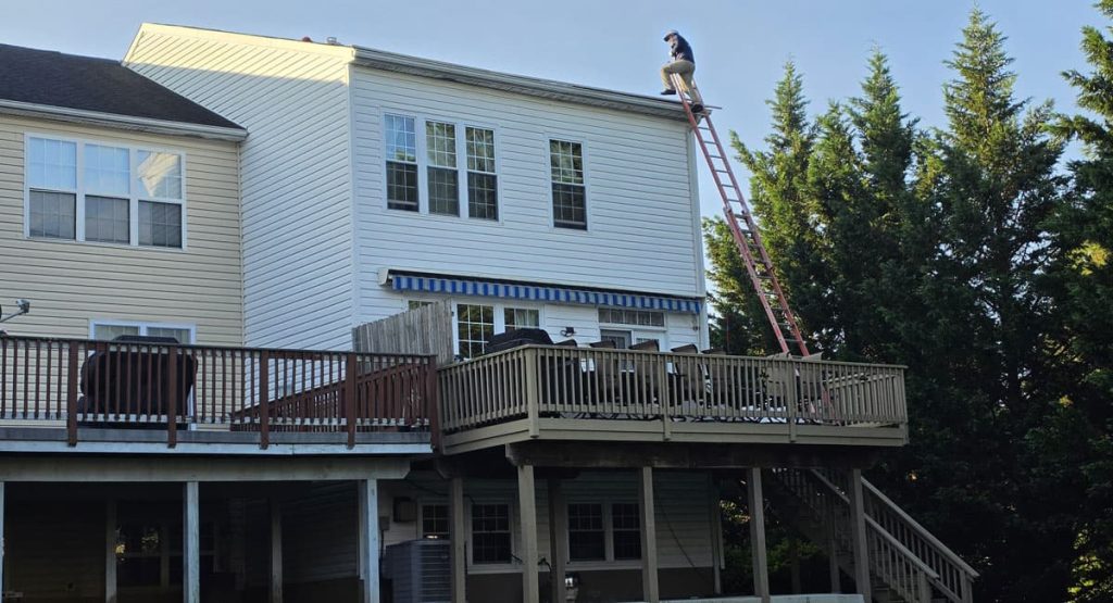 Person standing on a tall ladder leaning against the roof of a two-story house, doing maintenance on the upper edge, with a wooden deck in the foreground and trees nearby