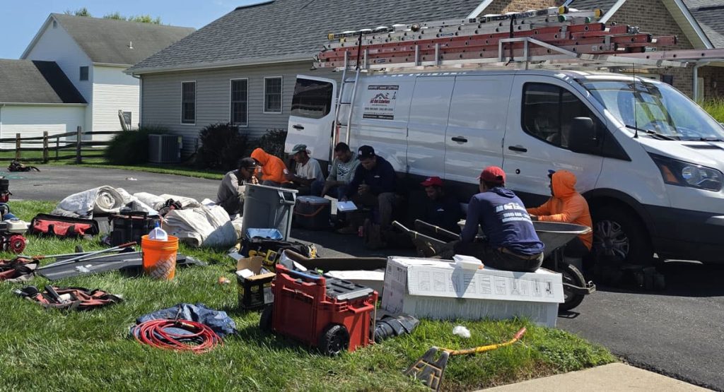 Crew of workers sitting on the curb and by a white van with ladders, taking a break amid tools and equipment for a residential repair job.