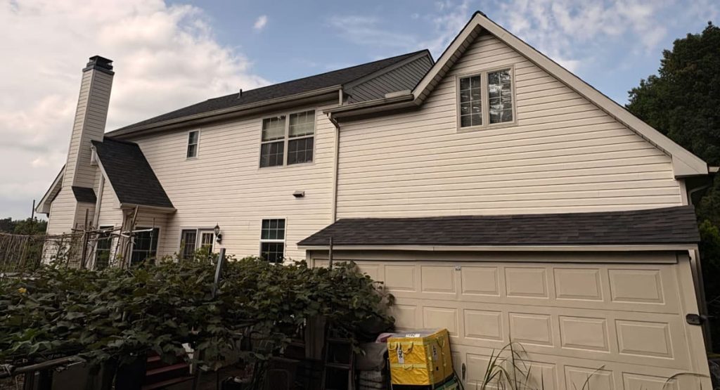 Two-story beige house with a front-facing garage, pitched roof, and a tall chimney on the left; plants in the foreground.